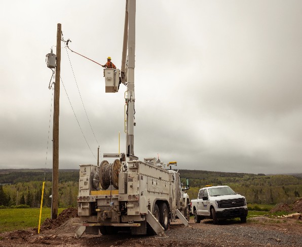 Utility worker in a bucket truck repairing overhead power lines on a wooden pole, with a second white utility truck parked nearby on a gravel road.